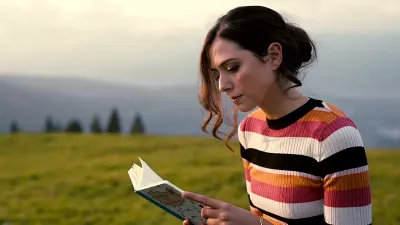 A young woman reading a book in a field. She is wearing a striped sweater and her hair is tied up.