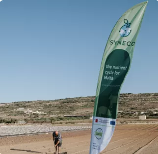 A man works in his field in front of a beach flag with the name of his project