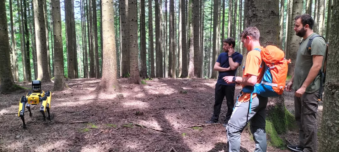3 people stand in a forest, one is demonstrating the use of robotics
