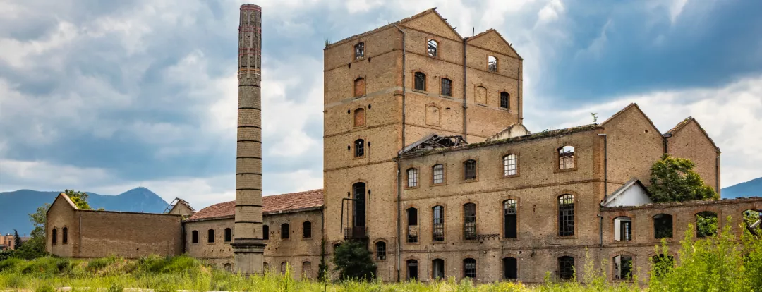 An old disused factory, abandoned and in ruins, with a smashed roof and a chimney. Tall weeds invade the building. Concept of economic bankruptcy. Cloudy sky. Italy, Foligno