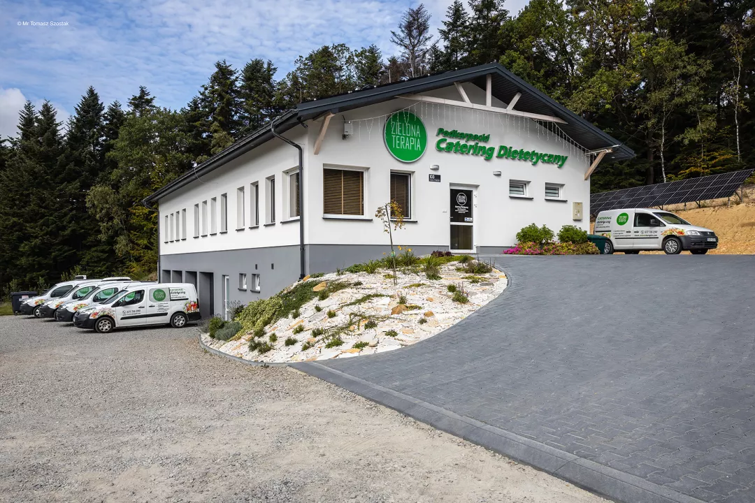 Modern white and gray building with 'Zielona Terapia' and 'Profesjonalny Catering Dietetyczny' signs, surrounded by landscaped plants, with delivery vans parked nearby