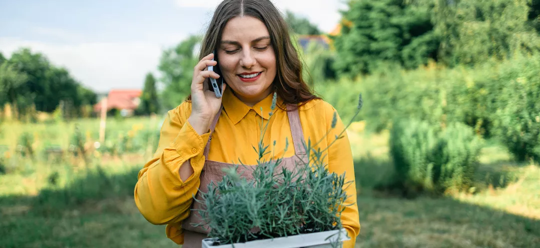 happy female Caucasian farmer talking on the smartphone and holding a plastic pot of lavender flower plants